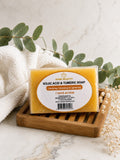 A rectangular bar of Nois Beauty Kojic Acid & Tumeric Soap in a clear plastic wrapper with a white label, displayed on a wooden soap dish with a white towel, eucalyptus leaves, and dried flowers.