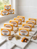 Multiple bars of Kojic Aloe & Turmeric Soap arranged on white marble tiles in a bright bathroom-like setting, with small props including dried flowers, twine, and turmeric roots.