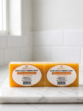 Two bars of Home Beauty Kojic Acid & Turmeric Soap resting on a white marble surface against a white tiled background.