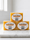 Three rectangular bars of Nourish Beauty Kojic Acid & Turmeric Soap arranged in a pyramid stack on a white marble surface with water droplets, against a bright, softly blurred background.